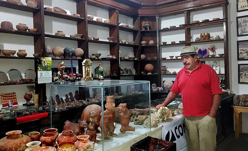 In a red polo shirt and khaki pants, a middle -aged Mexican man stands within a small museum full of archaeological pottery and sculptures, in some shelves and some glass events.