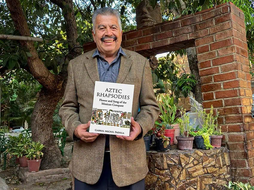 Author Gabriel Michael Stands in a small rock garden while holding her book,