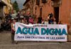Protest in a downtown street of San Cristobal de las Casas, Chiapas, with a line of people marching. At the front of the crowd, people are holding a white sign with black and agua-blue letters saying "We demand worthy water for San Cristobal de las Casas."