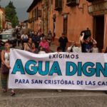 Protest in a downtown street of San Cristobal de las Casas, Chiapas, with a line of people marching. At the front of the crowd, people are holding a white sign with black and agua-blue letters saying "We demand worthy water for San Cristobal de las Casas."