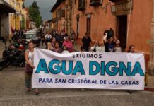 Protest in a downtown street of San Cristobal de las Casas, Chiapas, with a line of people marching. At the front of the crowd, people are holding a white sign with black and agua-blue letters saying "We demand worthy water for San Cristobal de las Casas."