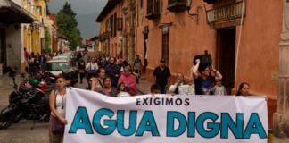 Protest in a downtown street of San Cristobal de las Casas, Chiapas, with a line of people marching. At the front of the crowd, people are holding a white sign with black and agua-blue letters saying "We demand worthy water for San Cristobal de las Casas."