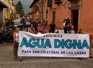 Protest in a downtown street of San Cristobal de las Casas, Chiapas, with a line of people marching. At the front of the crowd, people are holding a white sign with black and agua-blue letters saying "We demand worthy water for San Cristobal de las Casas."
