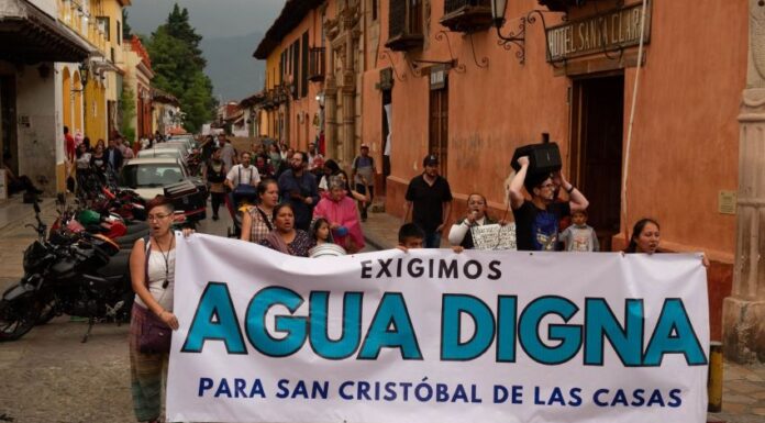 Protest in a downtown street of San Cristobal de las Casas, Chiapas, with a line of people marching. At the front of the crowd, people are holding a white sign with black and agua-blue letters saying "We demand worthy water for San Cristobal de las Casas."