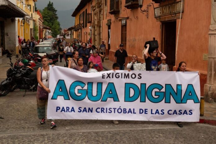 Protest in a downtown street of San Cristobal de las Casas, Chiapas, with a line of people marching. At the front of the crowd, people are holding a white sign with black and agua-blue letters saying 