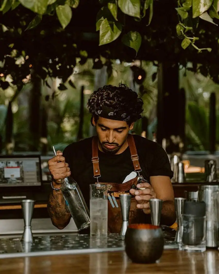 A Mexican Bartender is in the middle of mixing a black t-shirt on a black t-shirt, and a bandanna is in the middle of mixing the cocktail in a tall mirror in a tall mirror.