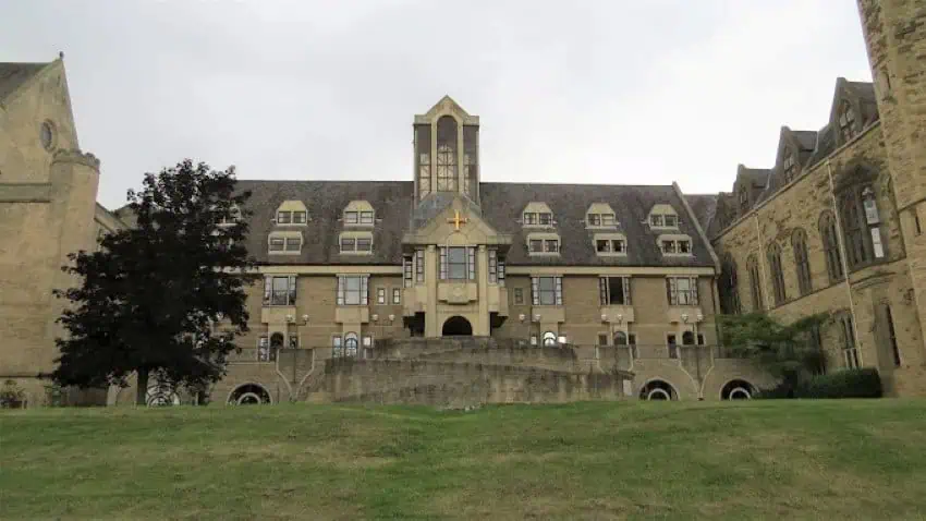 A hundreds of years old sandstone brick estate in Yorkshire with a small gold cross at the front entrance. At its front is an expanse of well-manicured lawn.