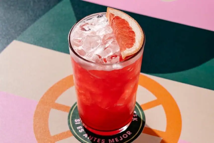 tall highball glass with a grapefruit colored drink. There is a grapefruit wedge resting atop the drink's ice cubes floating at the top of the glass.