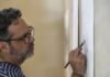 A man stands working on a pencil drawing mounted on a wall.