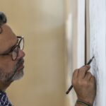 A man stands working on a pencil drawing mounted on a wall.