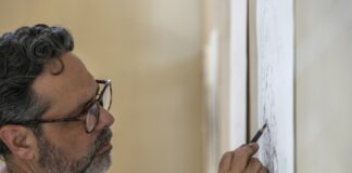 A man stands working on a pencil drawing mounted on a wall.
