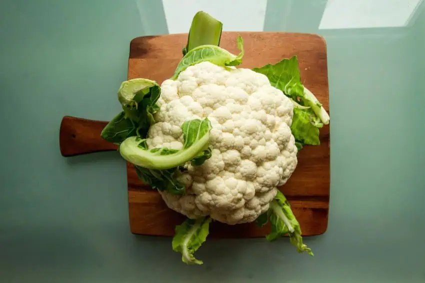 A whole cauliflower with green leaves still attached, sitting on a wooden cutting board on a green background.