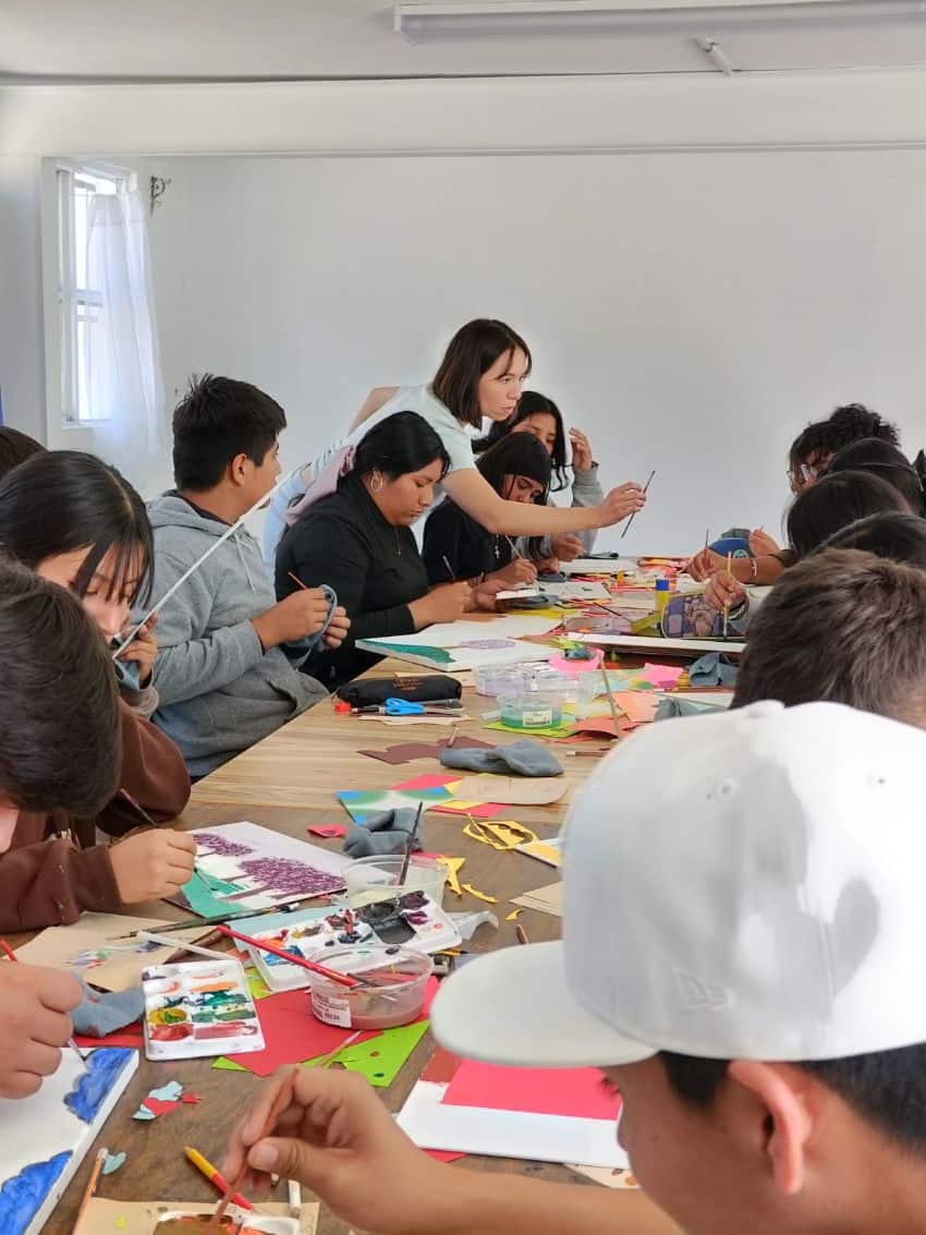 An art teacher holds an art paint brush towards a student on a big desk, where many adult students work in painting programs.