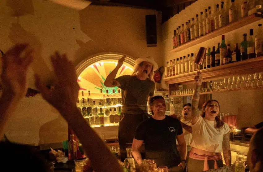 Bar staff and customers cheering and holding up a drink mixer in celebration.