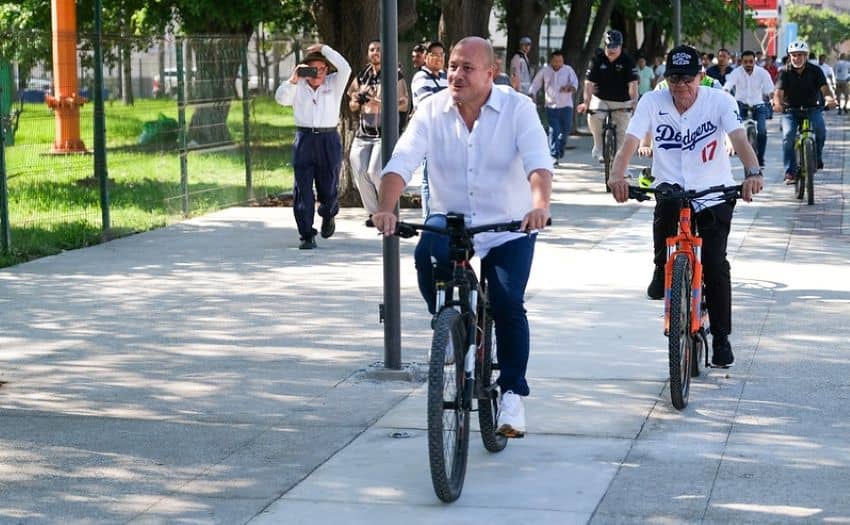 Jalisco's Governor Enrique Alfaro riding a bike on a new bike lane in Puerto Vallarta. He's in the lead wearing a white button down cotton shirt and black pants and white sneakers on a black mountain bike. Behind him at varying distances, are other bicyclists. In the background, a man in a dress shirt and black pants takes footage of him with a camera.