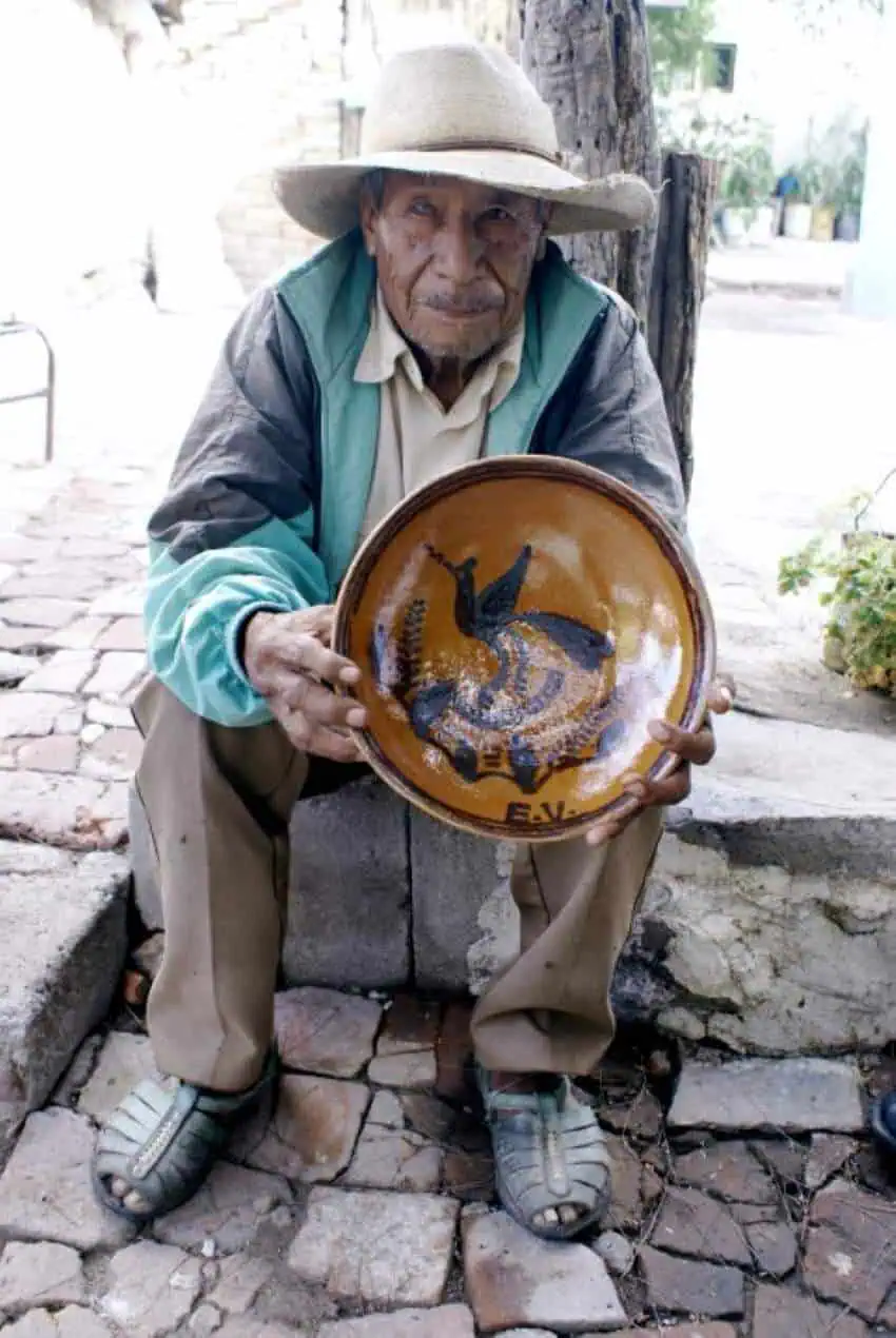 An elderly man wearing a hat on the edge of straw sits on the lower stone wall on the city street and holds a ceramic bowl in front of him.