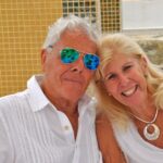 An elderly man and woman holding each other and posing for the camera outside against a Mexican tiled wall.