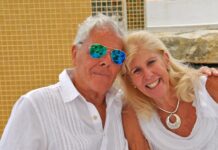 An elderly man and woman holding each other and posing for the camera outside against a Mexican tiled wall.