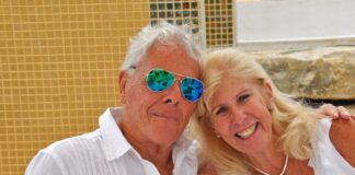 An elderly man and woman holding each other and posing for the camera outside against a Mexican tiled wall.
