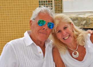 An elderly man and woman holding each other and posing for the camera outside against a Mexican tiled wall.