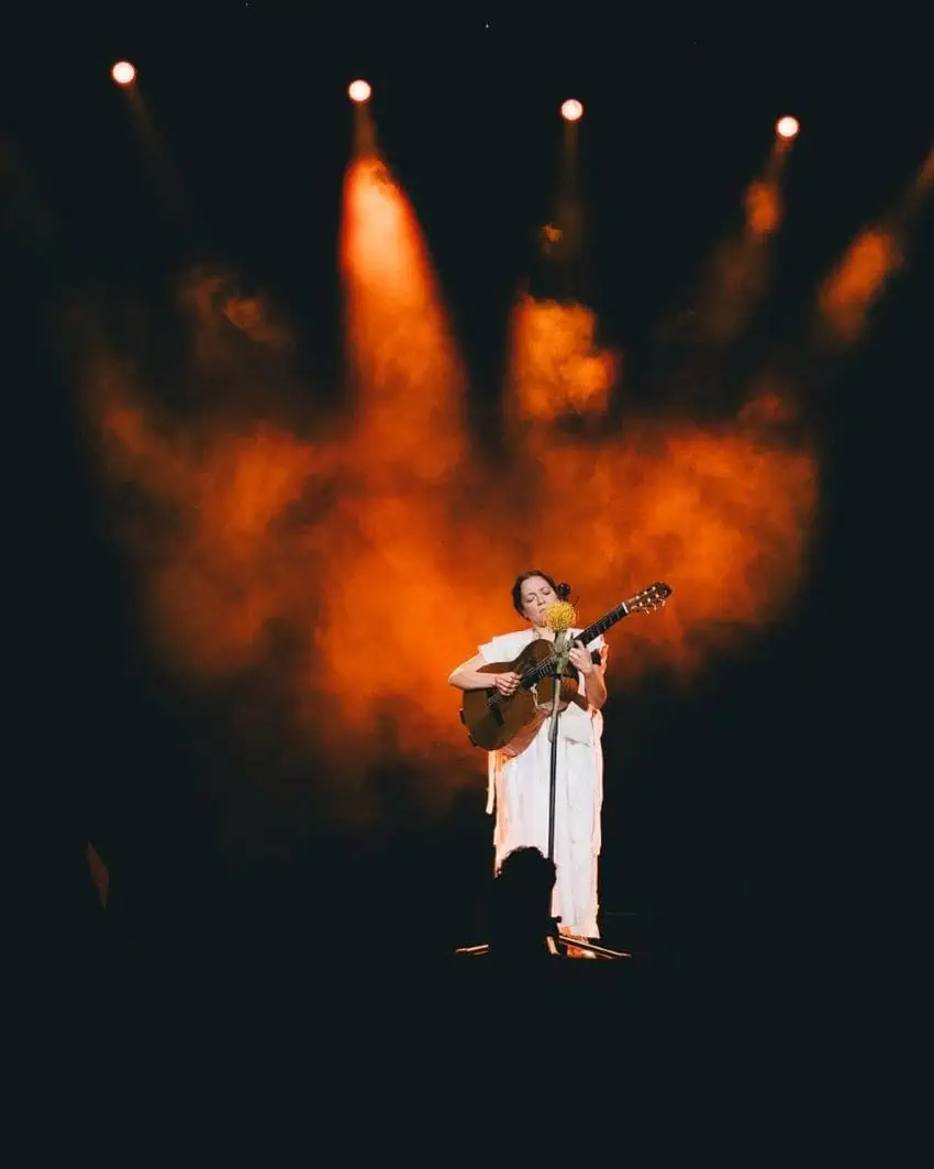 Natalia Lafourcade stands onstage at a performance on the Cancionera tour playing a guitar and dressed in white