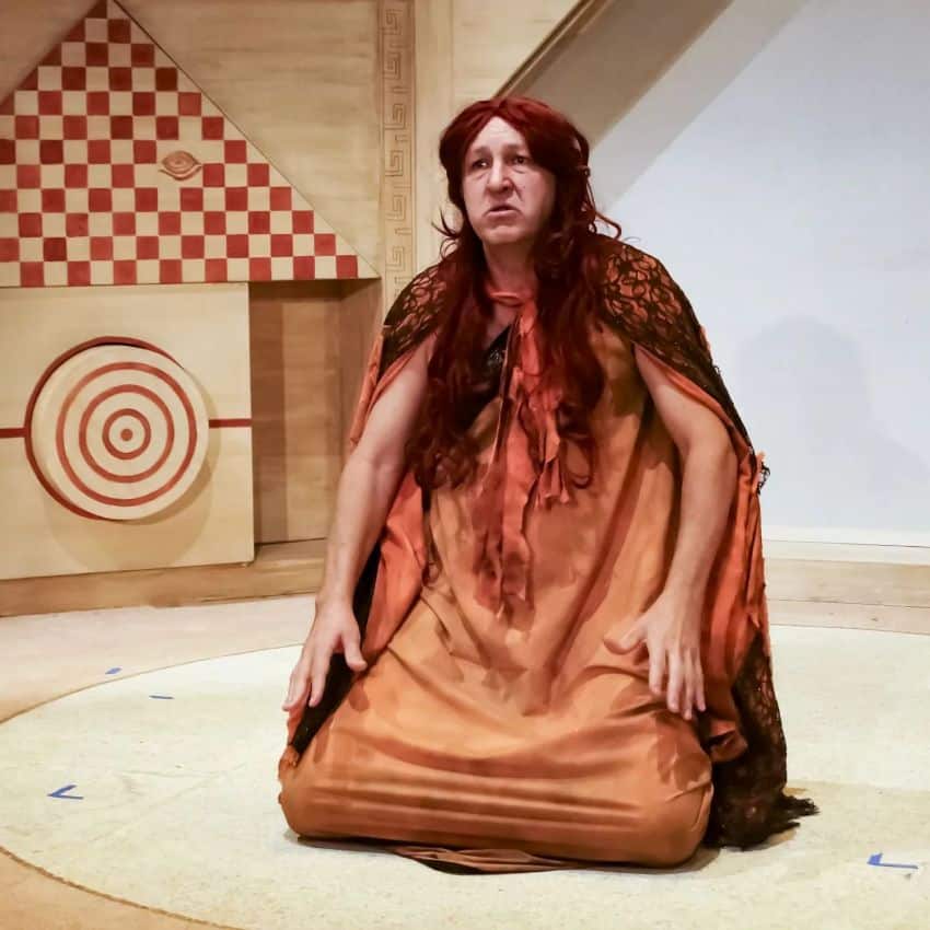 A middle aged woman in an orange full-length shift and a matching cape covered with black lace kneels on a bare stage. She looks outward at the audience with a chagrined expression