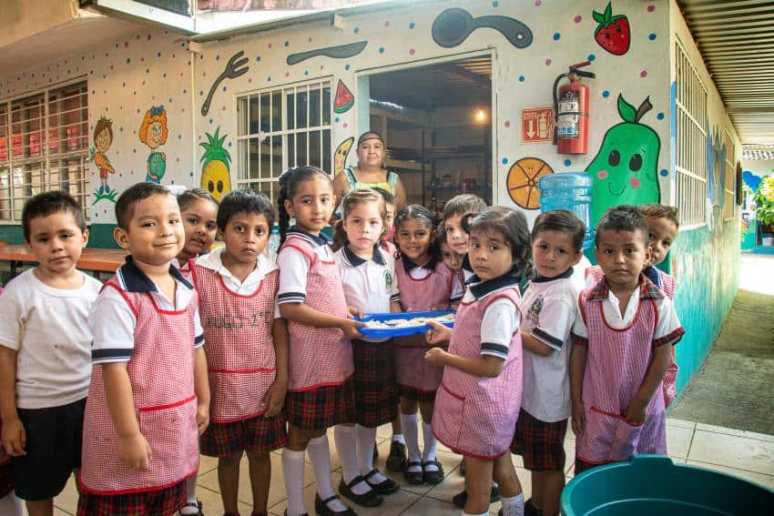 A group of schoolchildren in typical Mexican aprons holding a tray of food as they smile for the camera in their outdoor cafeteria, which has colorful images of fruit and cutlery. A cafeteria cook wearing a hairnet stands in the background looking on.