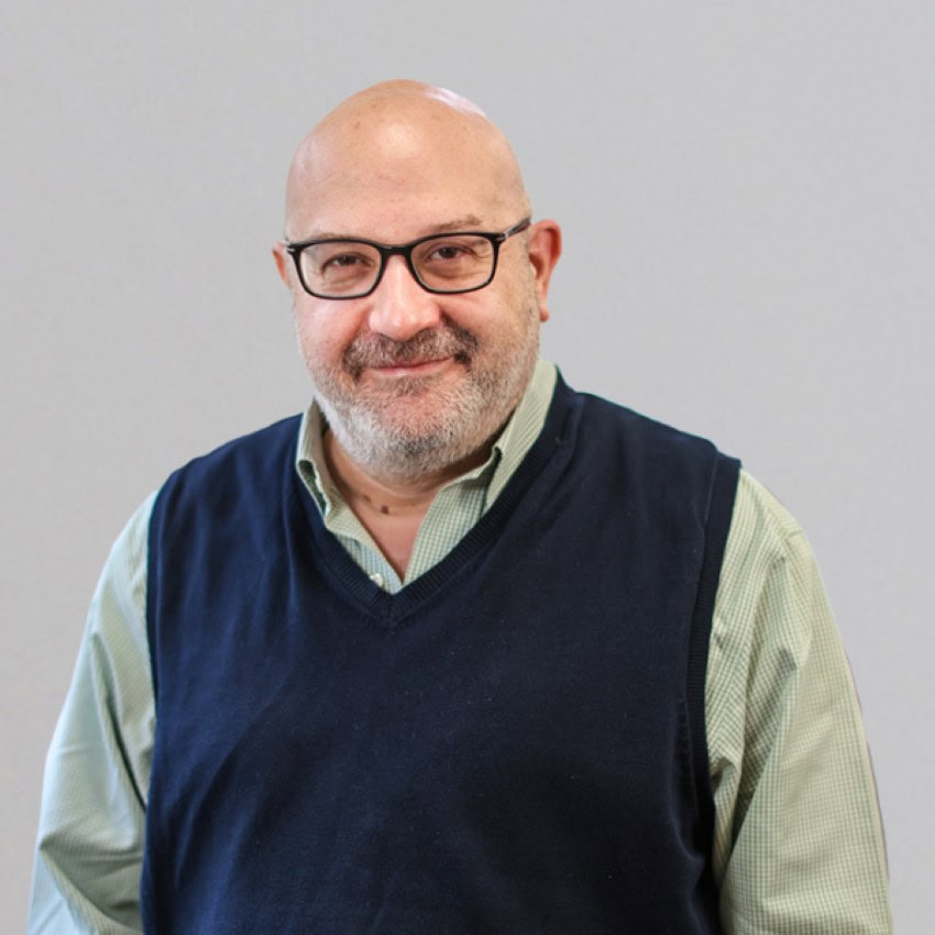 A bald man wearing glasses, a light green oxford and a navy blue sweater vest, smiles for the camera in a publicity photo with a bokeh background.