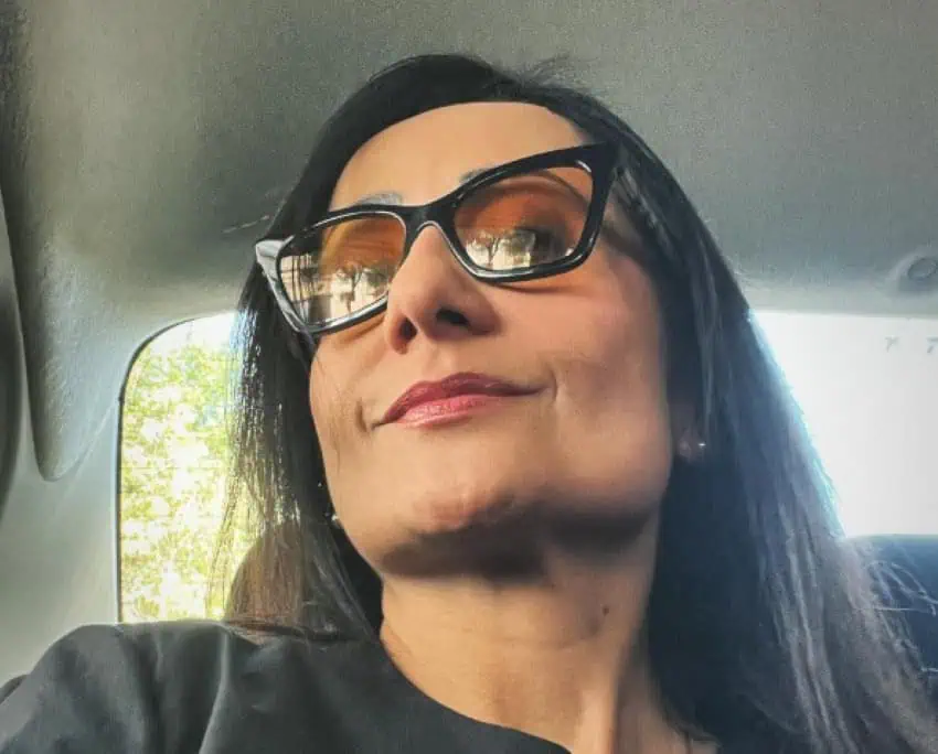 A woman with long hair and dense, black mirrors sits in a car.