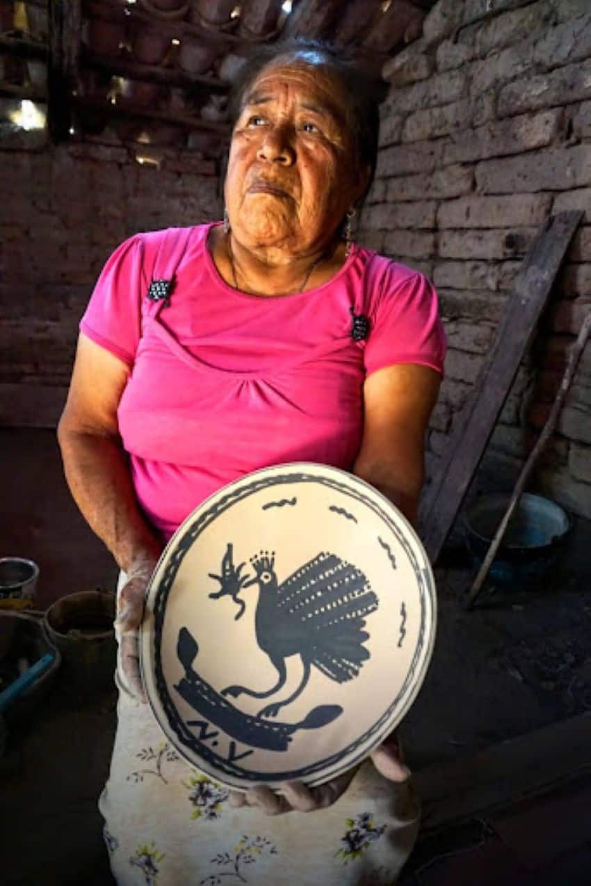 An elderly Mexican woman in a pink cotton short-sleeved shirt and a floral print skirt holds a large artisan pottery bowl she made. It's white with a large bird painted in the center in blue with a blue border around the edges. It has her intials paintedon it, N.V.