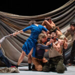 A stage in which six actors are in a performance, some kneeling, some standing, but all holding each other in some way. The man in the center of the group is looking upward as an actress holds up his chin