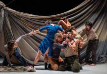 A stage in which six actors are in a performance, some kneeling, some standing, but all holding each other in some way. The man in the center of the group is looking upward as an actress holds up his chin