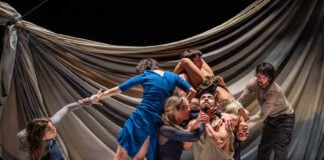 A stage in which six actors are in a performance, some kneeling, some standing, but all holding each other in some way. The man in the center of the group is looking upward as an actress holds up his chin