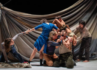 A stage in which six actors are in a performance, some kneeling, some standing, but all holding each other in some way. The man in the center of the group is looking upward as an actress holds up his chin