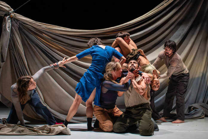 A stage in which six actors are in a performance, some kneeling, some standing, but all holding each other in some way. The man in the center of the group is looking upward as an actress holds up his chin