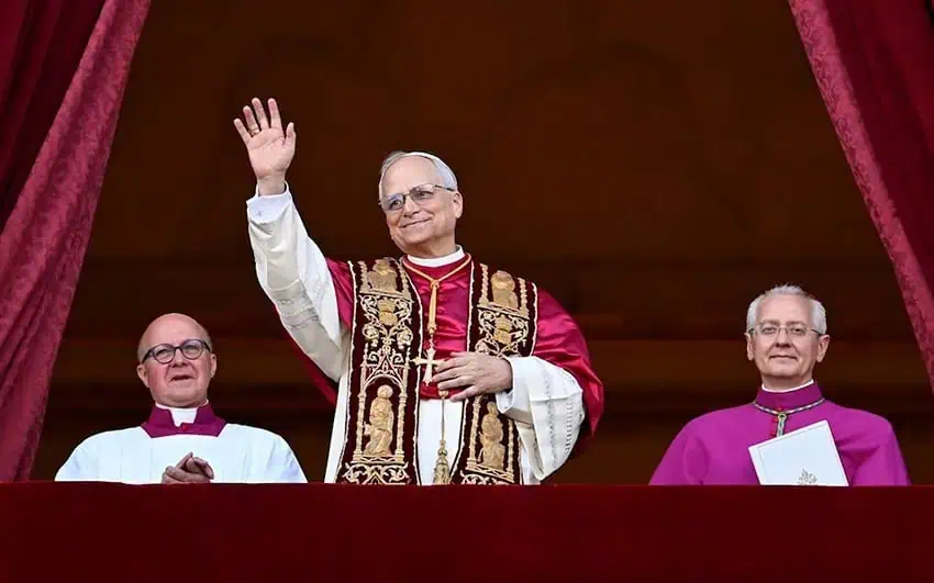 Pope Leo XIV waving to the public in St. Peter's Square after being shown to the public for the first time as the Catholic Church's new pope. He is wearing the Pope's official regalia. Beside him sit two church officials in vestments