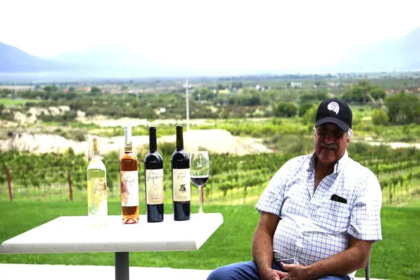 Jeans and a short sleeve button down the Cekerd shirt and a black baseball hat, sunglasses, wearing sunglasses, and looking at the camera when he sits on a small table with four bottles of wine from the same wine.