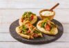 Three tlacoyos topped with peppers and onions, sitting on a serving platter made of volcanic rock. Also on this platter is a small wooden bowl of white rice with a matching wooden spoon for serving.