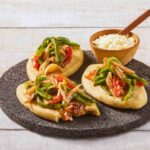 Three tlacoyos topped with peppers and onions, sitting on a serving platter made of volcanic rock. Also on this platter is a small wooden bowl of white rice with a matching wooden spoon for serving.