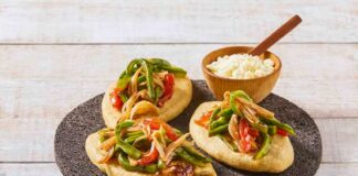 Three tlacoyos topped with peppers and onions, sitting on a serving platter made of volcanic rock. Also on this platter is a small wooden bowl of white rice with a matching wooden spoon for serving.