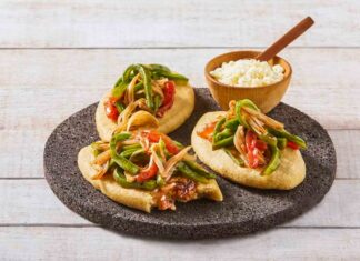 Three tlacoyos topped with peppers and onions, sitting on a serving platter made of volcanic rock. Also on this platter is a small wooden bowl of white rice with a matching wooden spoon for serving.
