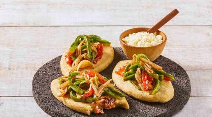 Three tlacoyos topped with peppers and onions, sitting on a serving platter made of volcanic rock. Also on this platter is a small wooden bowl of white rice with a matching wooden spoon for serving.