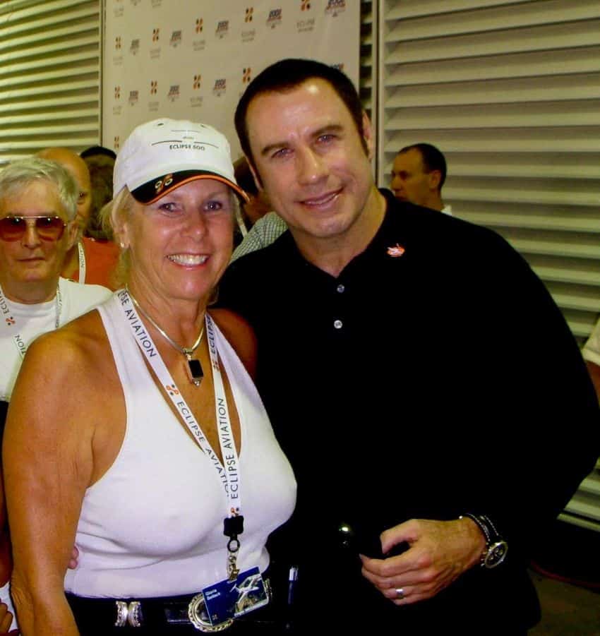 A blonde, middle aged woman poses with actor John Travolta for a selfie at an Eclipse Aviation event in the 1980s. Other guests look on behind them.