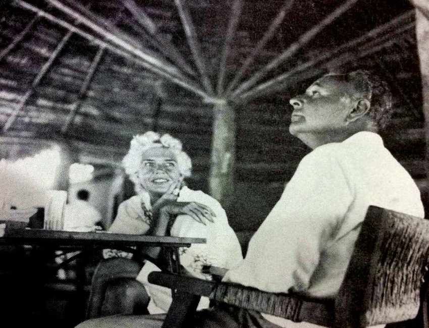 A achromatic and achromatic photograph of a middle-aged Mexican antheral successful a achromatic fastener down garment sitting astatine a array nether a operation with a palapa extortion and looking upward. Across from him is simply a middle-aged pistillate who is looking astatine him.