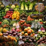 An open air market in a Spanish-speaking country where a wide variety of fruits and vegetables are on display with their prices. In the foreground, an old-fashioned hanging scale for food items can be seen.