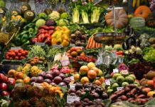 An open air market in a Spanish-speaking country where a wide variety of fruits and vegetables are on display with their prices. In the foreground, an old-fashioned hanging scale for food items can be seen.