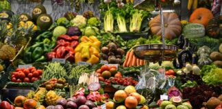 An open air market in a Spanish-speaking country where a wide variety of fruits and vegetables are on display with their prices. In the foreground, an old-fashioned hanging scale for food items can be seen.