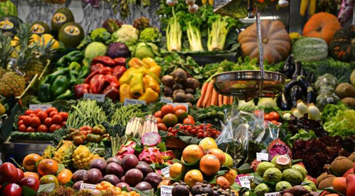 An open air market in a Spanish-speaking country where a wide variety of fruits and vegetables are on display with their prices. In the foreground, an old-fashioned hanging scale for food items can be seen.