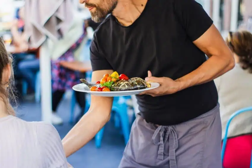 A man in a black tee shirt and wearing a mauve restaurant apron over his torso serves a plate of fish and colorful vegetables to an unseen customer in a restaurant environment.