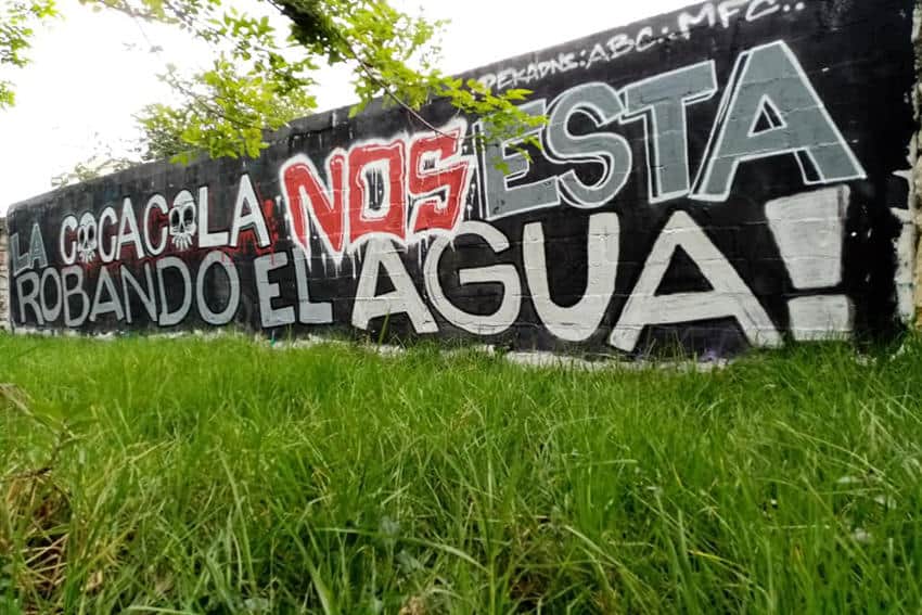 White and red graffitti on a black-painted brick wall in San Cristobal de Las Casas, Chiapas, that says in Spanish, "Coca Cola is robbing our water.